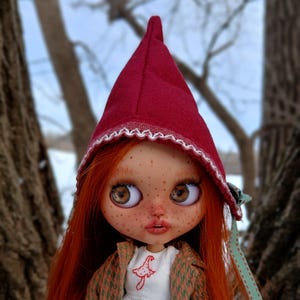 May include: A doll with long red hair, freckles, and large brown eyes, wearing a red hooded cap with white trim. The doll is dressed in a white dress and a green and brown patterned coat. A red mushroom is embroidered on the dress.