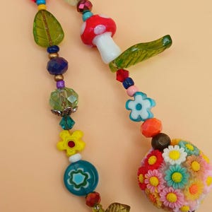 Könnte beinhalten: Eine skurrile Halskette mit bunten Glasperlen und Anhängern. Das Design umfasst einen roten Pilz, grüne Blätter und einen Blumenstrauß. Die Halskette hat einen roten Apfelanhänger und einen floralen Kugelpendant.