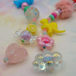 May include: A collection of colorful beaded charms and pendants. The charms include a yellow dinosaur, a pink flower, a pink heart, a lemon slice, and a candy-shaped charm. The beads are in various colors, including pink, blue, and clear.