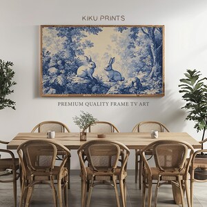 May include: A framed print of two white rabbits in a blue and white forest scene. The print is in a wooden frame and is hanging on a white wall. The text "KIKU PRINTS" is above the print and the text "PREMIUM QUALITY FRAME TV ART" is below the print.
