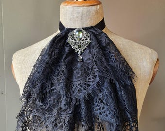 May include: A black lace jabot featuring a layered, ruffled design and a central silver-toned brooch with clear crystals and a dark teardrop-shaped pendant. The accessory is secured around the neck with a simple black fabric band.