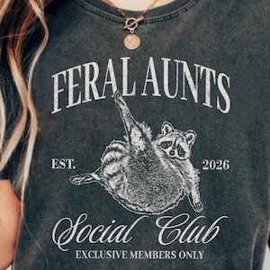 May include: Dark gray t-shirt with the text "FERAL AUNTS SOCIAL CLUB" and a raccoon graphic. The shirt includes the text "EST. 2026" and "EXCLUSIVE MEMBERS ONLY". The shirt is a vintage style.