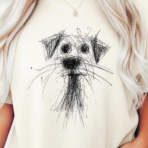 May include: Cream-colored t-shirt featuring a black and white sketch of a dog's face. The dog's eyes, nose, and whiskers are depicted with simple lines. The shirt has short sleeves and a relaxed fit. The brand logo is visible in the bottom right corner.