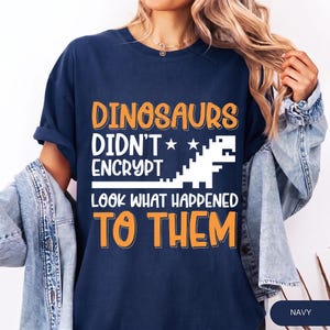 May include: A navy blue t-shirt featuring the orange and white text "DINOSAURS DIDN'T ENCRYPT LOOK WHAT HAPPENED TO THEM" alongside a white pixelated dinosaur graphic. The shirt is worn under a light blue denim jacket with visible metal buttons.