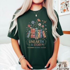 May include: Dark green t-shirt featuring a colorful graphic of stacked books, flowers, and butterflies. The text "UNEARTH A STORY" and "SUMMER READING 2026" are below the illustration. The shirt has short sleeves and a crew neck.