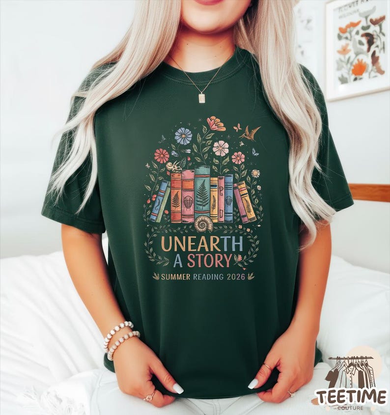 May include: Dark green t-shirt featuring a colorful graphic of stacked books, flowers, and butterflies. The text "UNEARTH A STORY" and "SUMMER READING 2026" are below the illustration. The shirt has short sleeves and a crew neck.