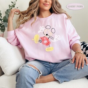 May include: A light pink sweatshirt with a neon yellow outline of Mickey Mouse in a playful pose. The sweatshirt has a crew neck and long sleeves.