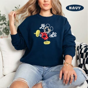 May include: A navy blue sweatshirt with a neon yellow and red outline of Mickey Mouse. The sweatshirt is worn by a person sitting on a white couch.