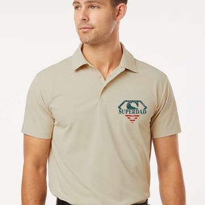 May include: Khaki polo shirt with a "Superdad" logo in a diamond shape on the chest. The logo features red, white, and blue colors. The shirt has short sleeves and a collared neckline.