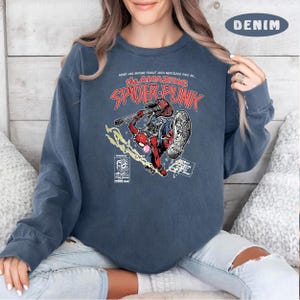 Puede incluir: Una sudadera azul con un gr&aacute;fico de un personaje de Spider-Man rojo y negro tocando una guitarra. El texto "The Amazing Spider-Punk" est&aacute; impreso en la sudadera.