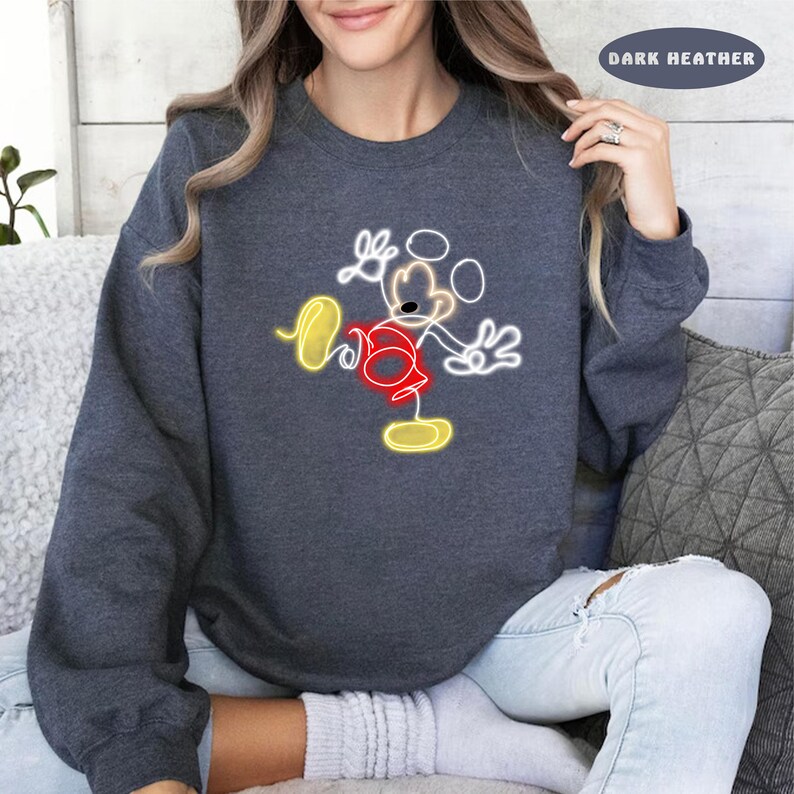 May include: A dark heather grey sweatshirt with a white outline of Mickey Mouse in a yellow and red color scheme. The text "DARK HEATHER" is visible in the top right corner of the image.