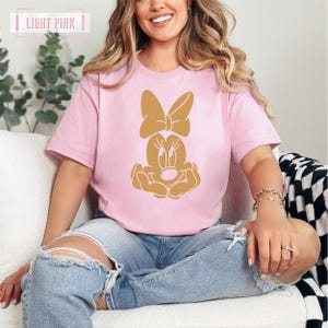 May include: A light pink t-shirt with a gold Minnie Mouse graphic. The graphic shows Minnie Mouse's face with her signature bow and hands clasped in front of her.