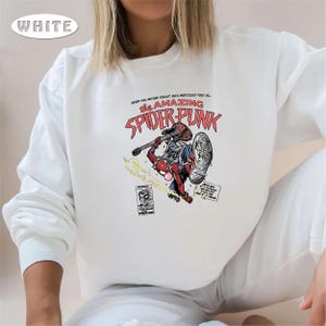 Puede incluir: Sudadera blanca de manga larga con un estampado gr&aacute;fico de un Spider-Man de dibujos animados tocando la guitarra. El texto "The Amazing Spider-Punk" est&aacute; impreso en rojo sobre la imagen.