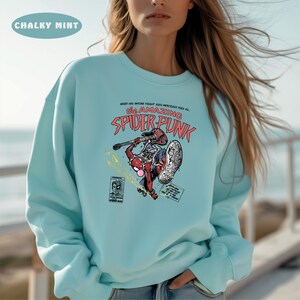 Puede incluir: Una sudadera de color azul claro con un estampado gr&aacute;fico de un superh&eacute;roe de dibujos animados tocando la guitarra. El texto en el gr&aacute;fico dice "The Amazing Spider-Punk".