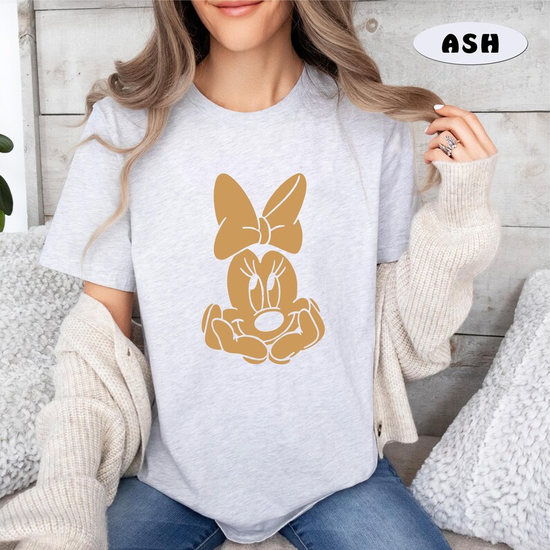 May include: A light gray t-shirt with a brown Minnie Mouse graphic. The graphic shows Minnie Mouse with her signature bow and a playful expression.