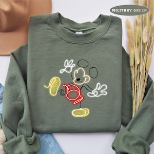 May include: A green sweatshirt with a white outline of Mickey Mouse in a neon yellow and red color scheme. The text "MILITARY GREEN" is in the top right corner of the image.