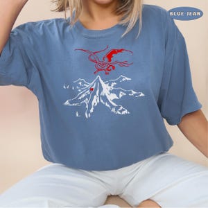 May include: A blue jean t-shirt with a white graphic of a mountain and a red dragon flying above it. The text "BLUE JEAN" is in the top right corner of the image.