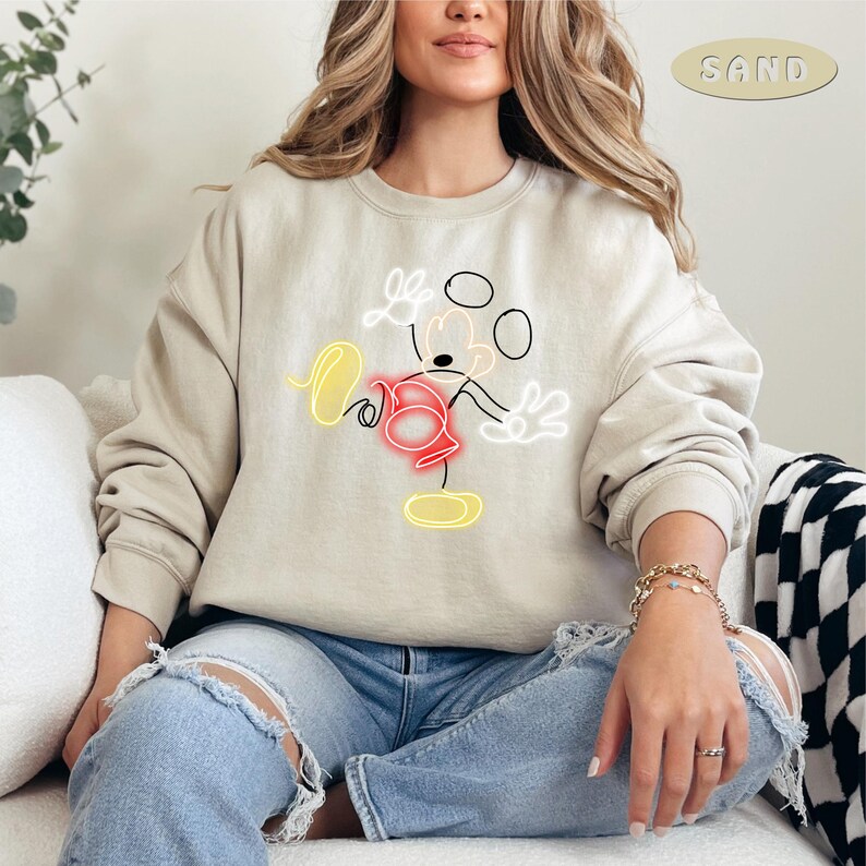 May include: A beige sweatshirt with a black and white outline of Mickey Mouse in a neon yellow and red color scheme. The text "SAND" is in the upper right corner of the image.