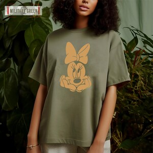 May include: A green t-shirt with a cartoon image of Minnie Mouse in orange. The image shows Minnie Mouse with her hands clasped in front of her.