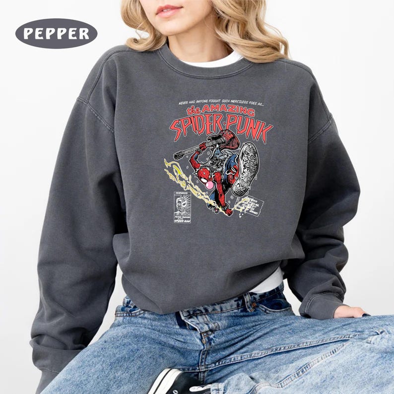 Puede incluir: Una sudadera gris oscuro con un estampado gr&aacute;fico de un superh&eacute;roe de dibujos animados sosteniendo una guitarra. El texto "The Amazing Spider-Punk" est&aacute; impreso en rojo y negro en la parte delantera de la sudadera.