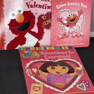 May include: A collection of children's Valentine's Day books featuring Elmo from Sesame Street and Dora the Explorer. The books are primarily pink and red, with illustrations of the characters and hearts. Titles include "My Fuzzy Valentine" and "Elmo Loves You."