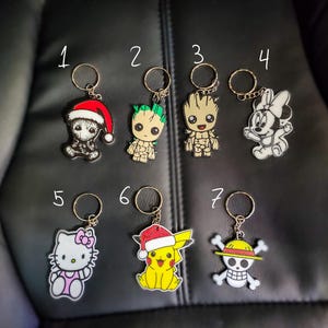 Peut inclure: Sept porte-clés différents représentant divers personnages de dessins animés. Les porte-clés sont numérotés de 1 à 7. Le porte-clés 1 représente un personnage de Père Noël vert et rouge. Le porte-clés 2 représente un personnage de Groot vert et marron. Le porte-clés 3 représente un personnage de Groot marron et vert. Le porte-clés 4 représente un personnage de Mickey Mouse blanc et noir. Le porte-clés 5 représente un personnage de Hello Kitty rose. Le porte-clés 6 représente un personnage de Pikachu jaune portant un bonnet de Père Noël rouge. Le porte-clés 7 représente un personnage de crâne blanc et noir portant un chapeau de paille.
