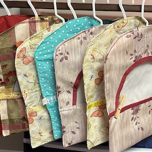 May include: A collection of six garment bags hanging on white hangers. The bags feature various patterns, including solid red, plaid, polka dots, and floral designs. Each bag has a unique design and color scheme, offering a variety of styles.