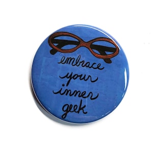 May include: A round, blue button with the words "embrace your inner geek" in black script. The button features a pair of brown-rimmed glasses at the top. The background is a solid blue color.