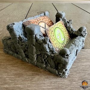 May include: A gray, 3D-printed card holder with a rocky, fortress-like design. The holder features skull accents and holds several game cards with green and brown designs. The Townbuilder 3D logo is visible in the bottom right corner.