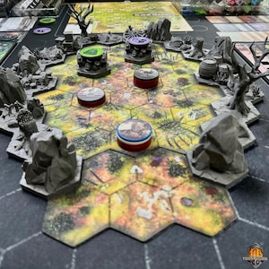 May include: A tabletop board game setup with a hexagonal game board. The board features a landscape design with various terrain elements. Gray miniature structures and tokens are placed on the board, with a dark background.
