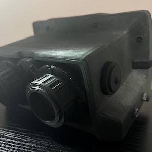May include: A dark green, 3D printed device with two black, round lenses and a black, round knob on the side.