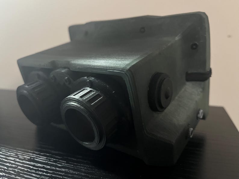 May include: A dark green, 3D printed device with two black, round lenses and a black, round knob on the side.