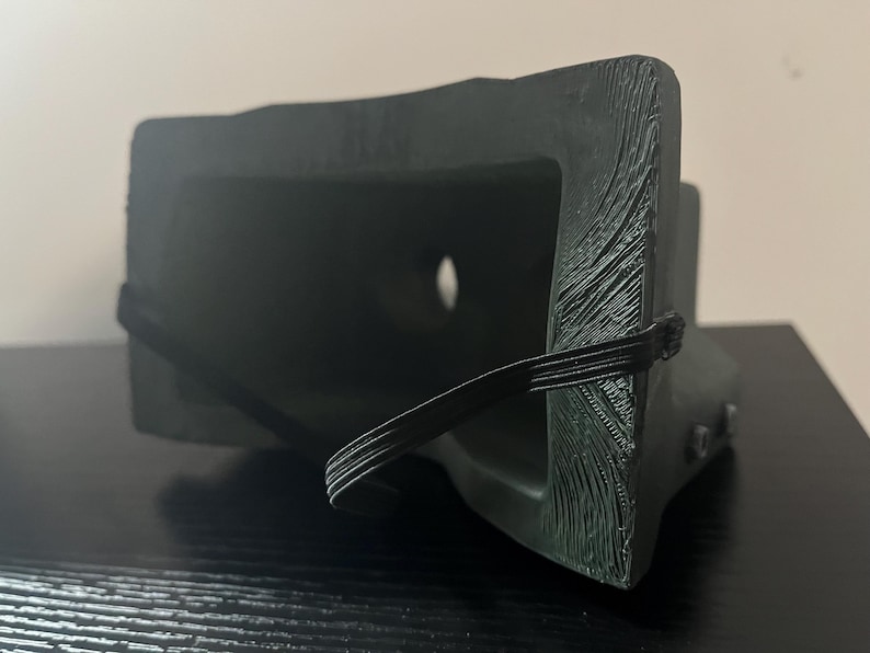 May include: A black plastic VR headset with an elastic strap. The headset has a textured surface and a small hole in the front.