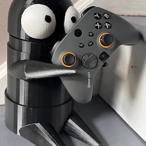 May include: A black 3D printed figurine of a character with large eyes holding a grey Scuf gaming controller with orange buttons.