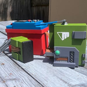 May include: A collection of colorful, 3D-printed objects. A red box with a blue top and black buttons sits next to a green box with a gray panel and a green button. A smaller green and black box is also present, along with a silver metal piece.