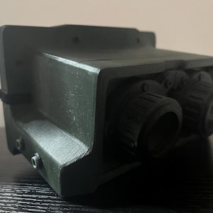 May include: A black and green plastic device with two round lenses. The device has a textured surface and appears to be a type of night vision equipment.