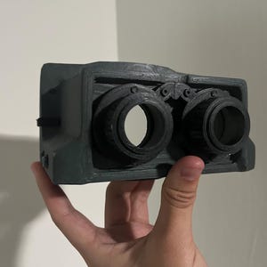 May include: A black 3D printed camera with two lenses. The camera has a textured surface and a detailed design.