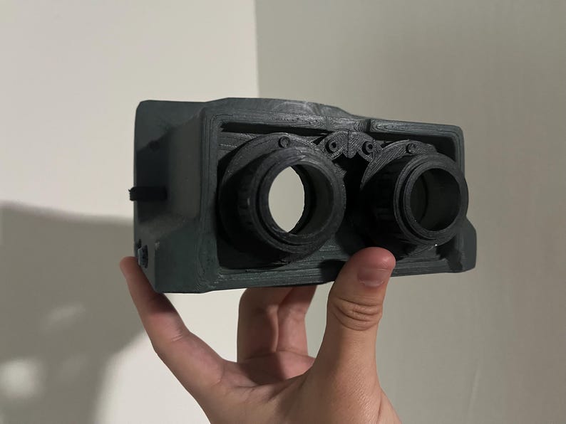 May include: A black 3D printed camera with two lenses. The camera has a textured surface and a detailed design.