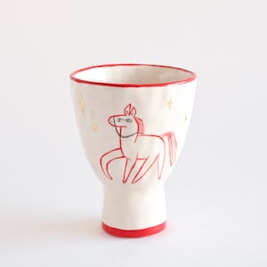 Może przedstawiać: Biały ceramiczny kubek z czerwonym obramowaniem i podstawą. Kubek posiada czerwony konturowy rysunek konia i złote gwiazdki. Kubek ma około 13 cm wysokości.