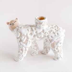 May include: A ceramic sculpture of a bear-like creature with a cream-colored body and white, textured accents. The bear has a small, tan-colored head with painted facial features and a cylindrical opening on its back.