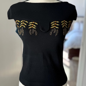 May include: Black knit top with a wide neckline and short sleeves. The top features an embroidered design in gold and brown, with a scalloped detail across the chest. The garment is displayed on a white mannequin.