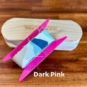 May include: A dark pink fabric ribbon winder with a wooden base. The ribbon is patterned with blue and white abstract shapes. The winder is labeled "Quilter's Apprentice".