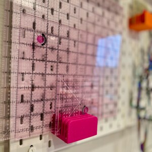 May include: A pink plastic ruler holder with a clear plastic ruler attached to it. The ruler holder is mounted on a white pegboard.