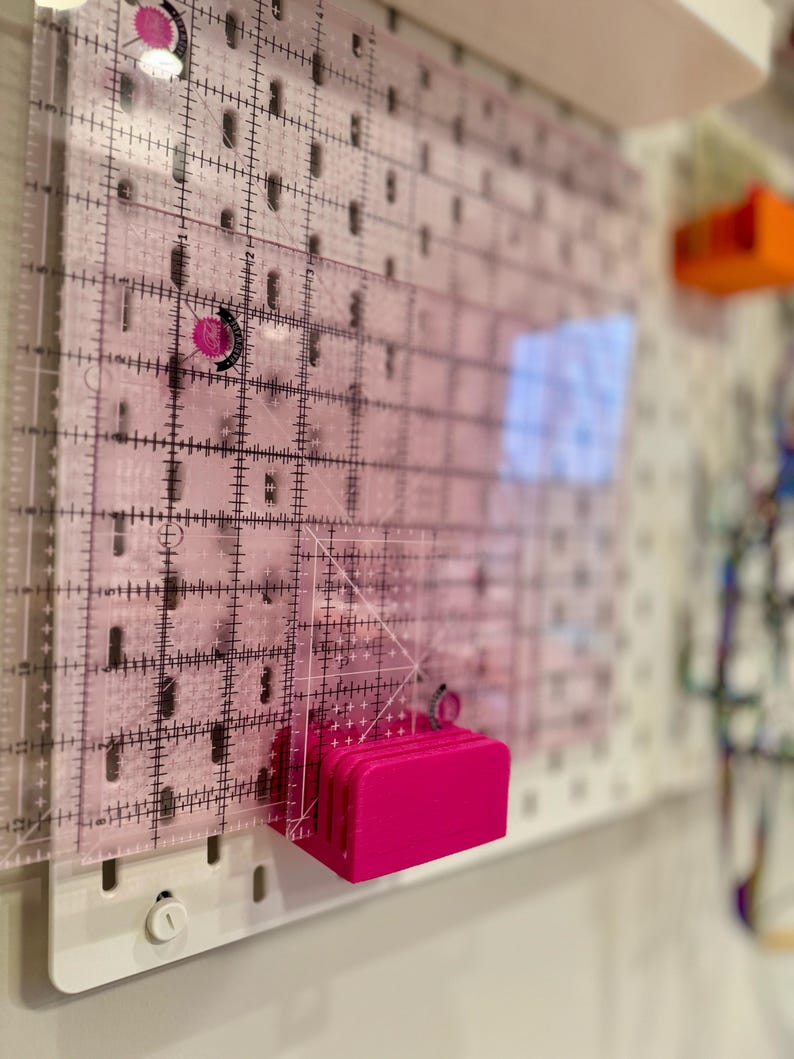 May include: A pink plastic ruler holder with a clear plastic ruler attached to it. The ruler holder is mounted on a white pegboard.