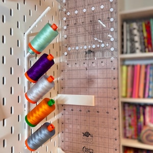 May include: A white pegboard with a metal thread spool holder and a clear quilting ruler. The holder contains spools of thread in green, purple, silver, orange, and gray. The ruler has grid lines and measurements.