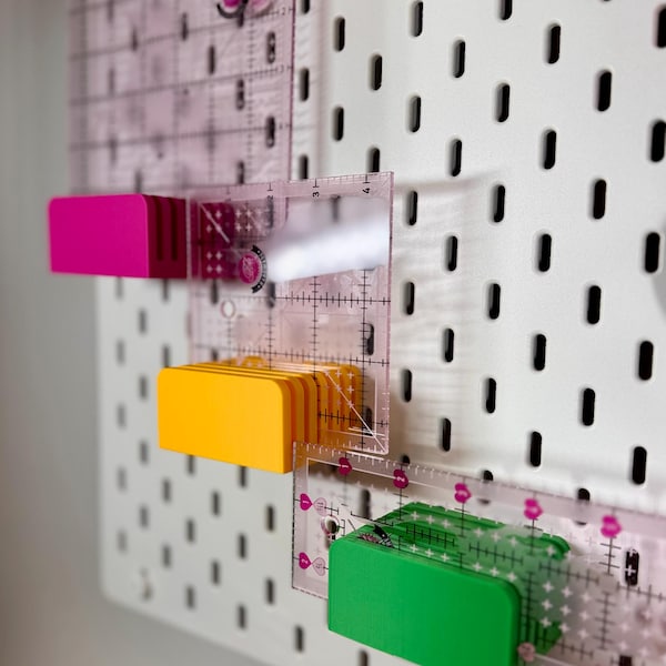 May include: Three clear acrylic quilting rulers with pink, yellow, and green rectangular plastic holders attached to a white pegboard. Each ruler features black grid markings and measurements in inches, designed for precise fabric cutting and alignment.
