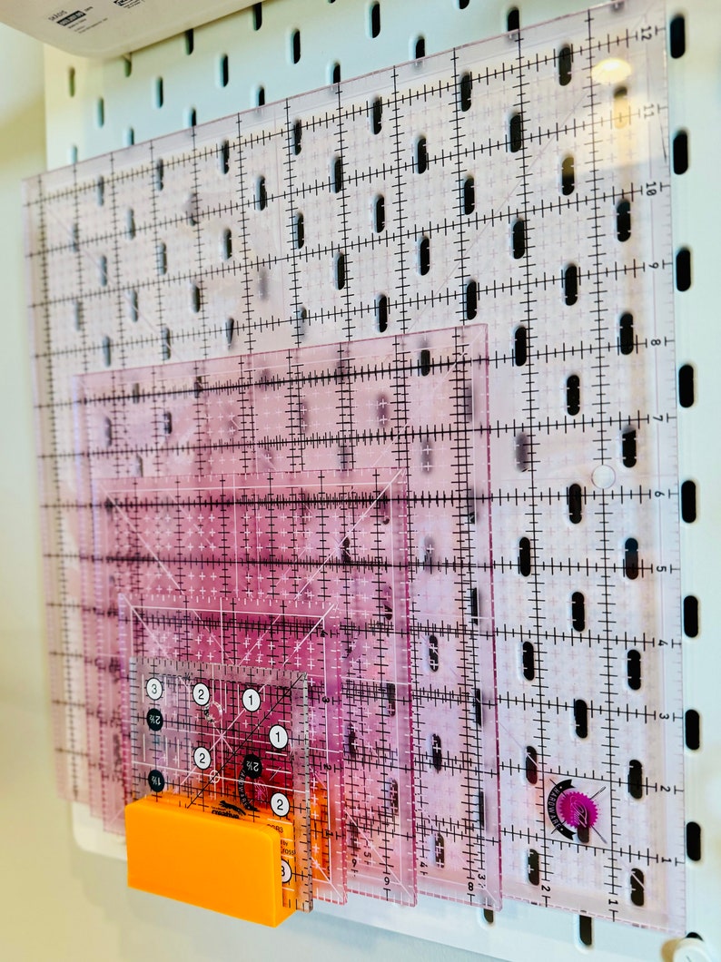 May include: A set of clear plastic quilting rulers with grid markings and inch measurements. The rulers are stacked and held in place by an orange magnetic clip.