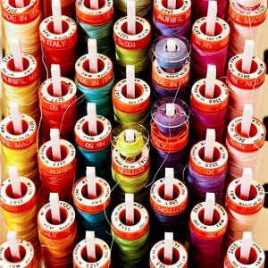 May include: A collection of colorful spools of thread, each with a white center and a label that reads "MADE IN ITALY." The threads are in various colors, including red, yellow, green, blue, and pink. The spools are arranged closely together.