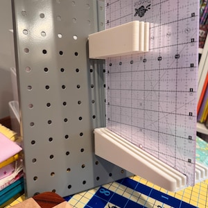 May include: A gray pegboard organizer featuring two white 3D-printed plastic brackets attached to the front. A clear acrylic quilting ruler with black grid markings and the text Poppie Cotton is slotted into the white brackets for vertical storage.