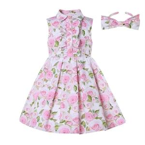 May include: A sleeveless white dress with a pink rose pattern, a ruffled front, and a matching bow headband. The dress has a collared neckline and a pleated skirt, perfect for a special occasion.